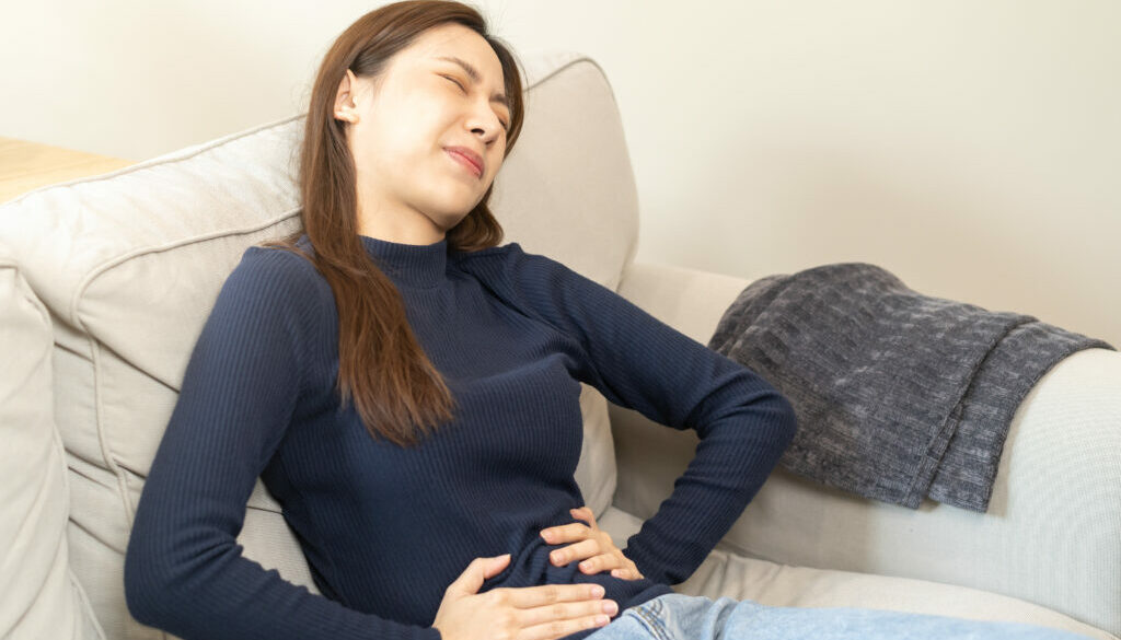 A Young Asian Woman Sitting on a Couch Holding Her Stomach in Pain Suffering From Types of Peptic Ulcers