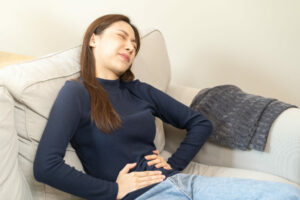 A Young Asian Woman Sitting on a Couch Holding Her Stomach in Pain Suffering From Types of Peptic Ulcers