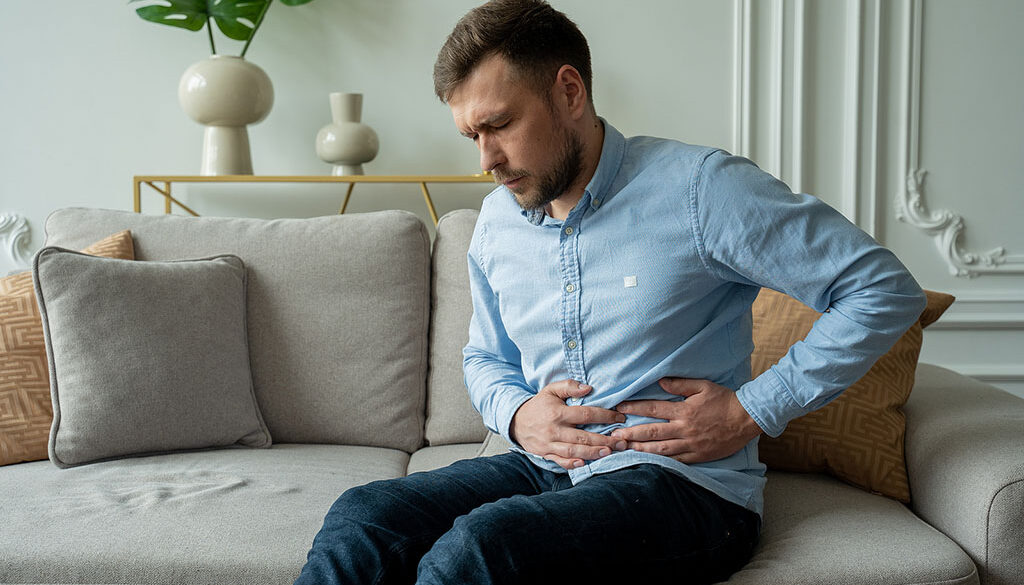 A White Man Sitting on a Couch Holding His Abdomen in Pain 7 Symptoms of Pancreas Problems