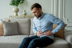 A White Man Sitting on a Couch Holding His Abdomen in Pain 7 Symptoms of Pancreas Problems