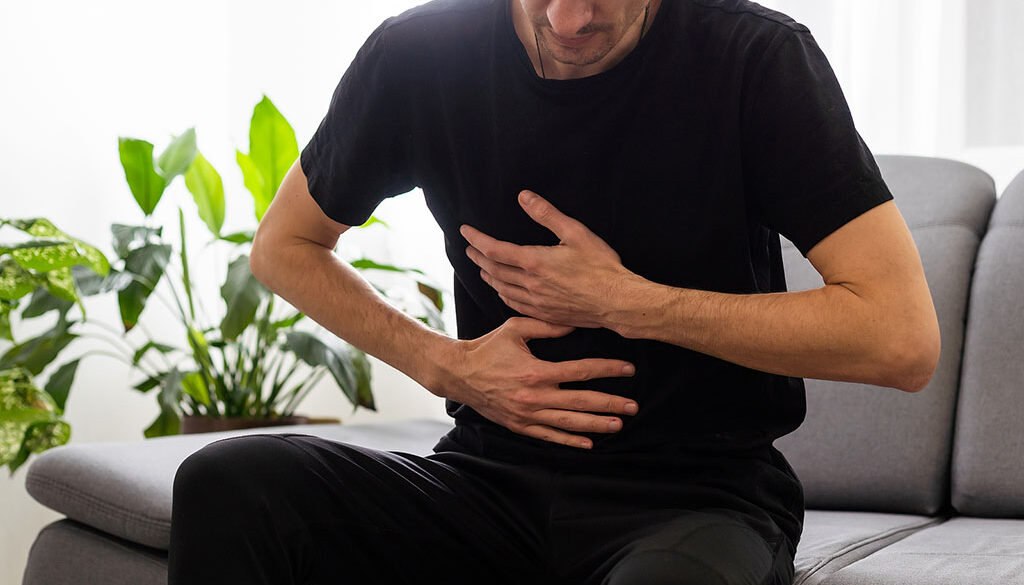 A Man Sitting On A Couch Holding His Upper Abdominal Area Can Gallstones Cause Acid Reflux