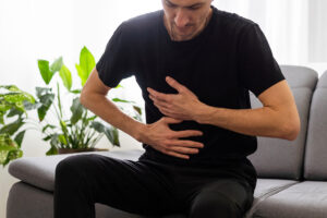 A Man Sitting On A Couch Holding His Upper Abdominal Area Can Gallstones Cause Acid Reflux