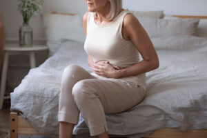 An Older Woman Sitting On The Edge Of A Bed Holding Her Stomach In Pain Gastritis In Women